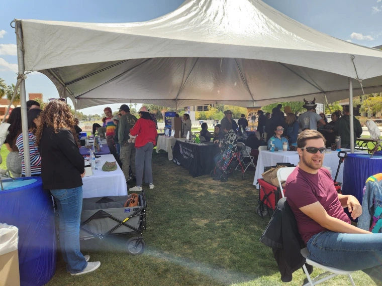 Another wide shot of the tabling area at Neurodiversity Day. The tables are on a grassy area under a large tent. Attendees are talking with the various organization representatives at their respective tables.