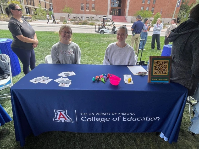 University of Arizona College of Education representatives sitting at their table. The table has a QR code, assorted fliers, and a few balloons