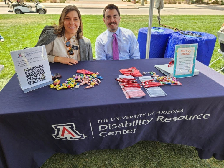 The University of Arizona Disability Resource Center representatives sitting at their table. The table has fliers, a QR code, and some assorted candies.