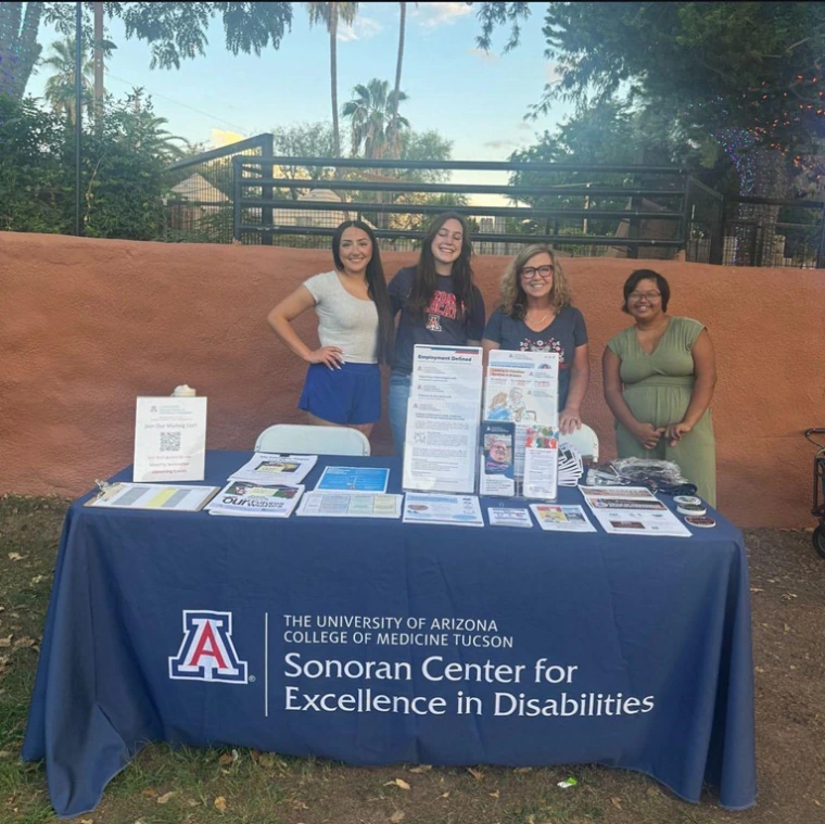 Sonoran Center staff standing and smiling behind a table with a variety of fliers, sign-up sheets, and other resources