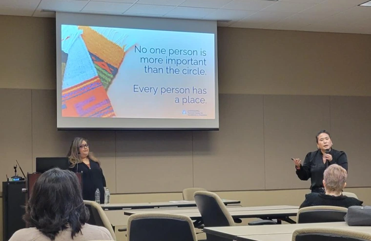 Rosa Rojas Franco and Mỹ Lê Sandaine giving their presentation. Mỹ Lê is holding a microphone and gesturing to a slide that reads "No one person is more important than the circle. Everyone has their place". 