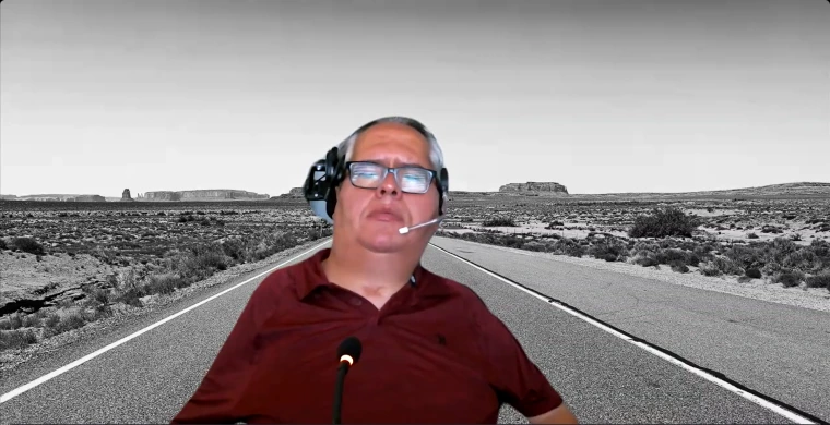 George Garcia opens the conference with introductions. He is wearing a red shirt and headphones, and is superimposed in front of a black and white desert background