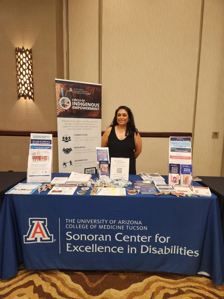 Celina Urquidez stands behind the Sonoran Center table at the AADMD conference. The table has a variety of pamphlets other resources displayed. 