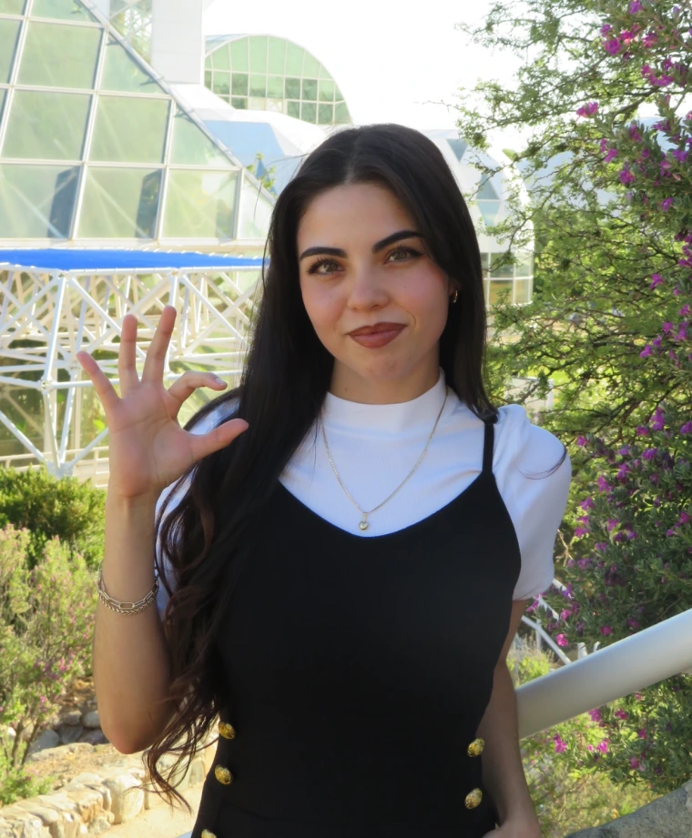 Subject is in the center of the picture, smiling and holding up her right arm, doing the "Wildcat" symbol (pinkie, ring and middle finger spread open, pointer and thumb curved into a "C" shape). Subject is wearing a plain white short sleeve, with a black jumpsuit over it. Subject is standing in front of Biosphere 2.