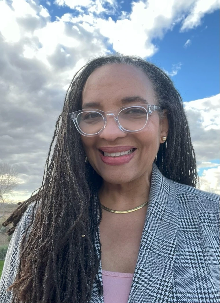Woman wearing a blue plaid blazer and pink shirt, she has long box braids wearing clear rimmed glasses