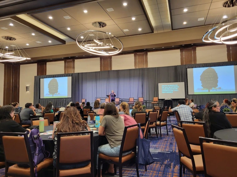 Rachel Simon delivers the closing remarks on the main stage at IHD. She is standing at a podium and gesturing to the crowd, next to a screen showing the cover of her book, "Riding the Bus with My Sister"
