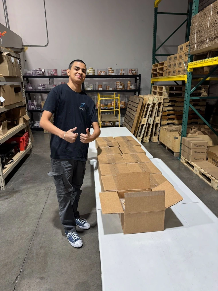 An employee participant stands next to sealed cardboard boxes and gives a double thumbs-up