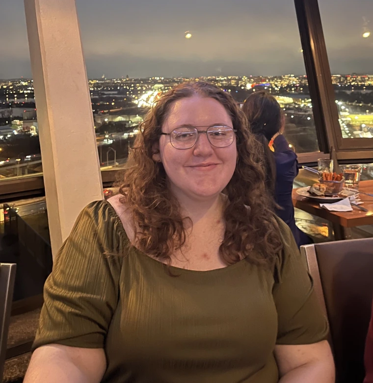 A woman sitting in front of a skyline view wearing a dark green shirt. She has mid length curly red hair and has brown glasses on. She sits at a dining table with a smile looking towards the camera.
