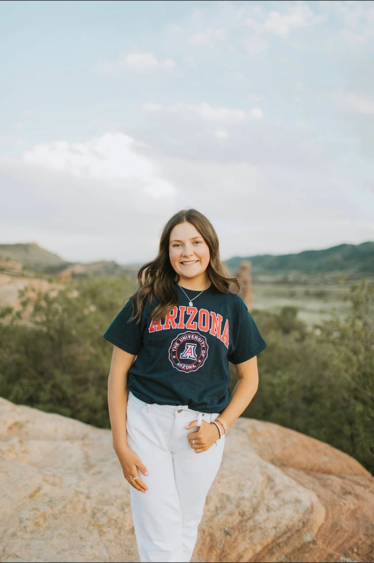 young woman wearing a blue U of A shirt and is smiling at the camera with a mountains background