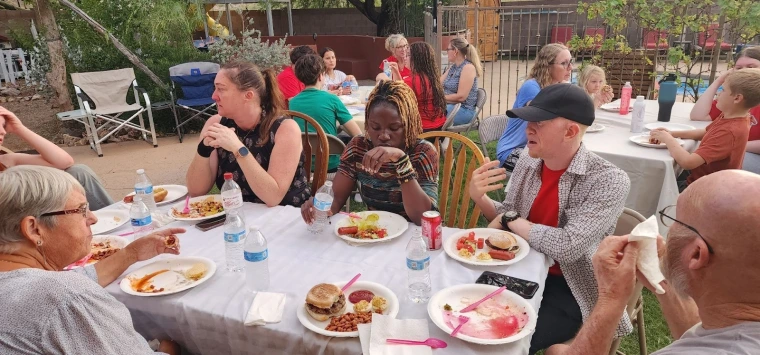 Mai and Kelvin sitting at a table and talking with other guests at a backyard barbeque on a clear day