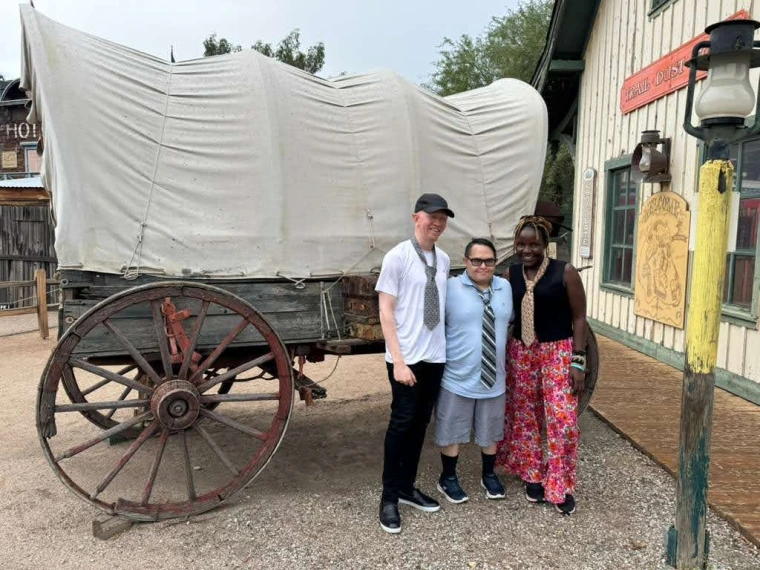 Kelvin, Gabe, and Mai standing outside an old-west style building in front of a covered wagon, all wearing novelty ties