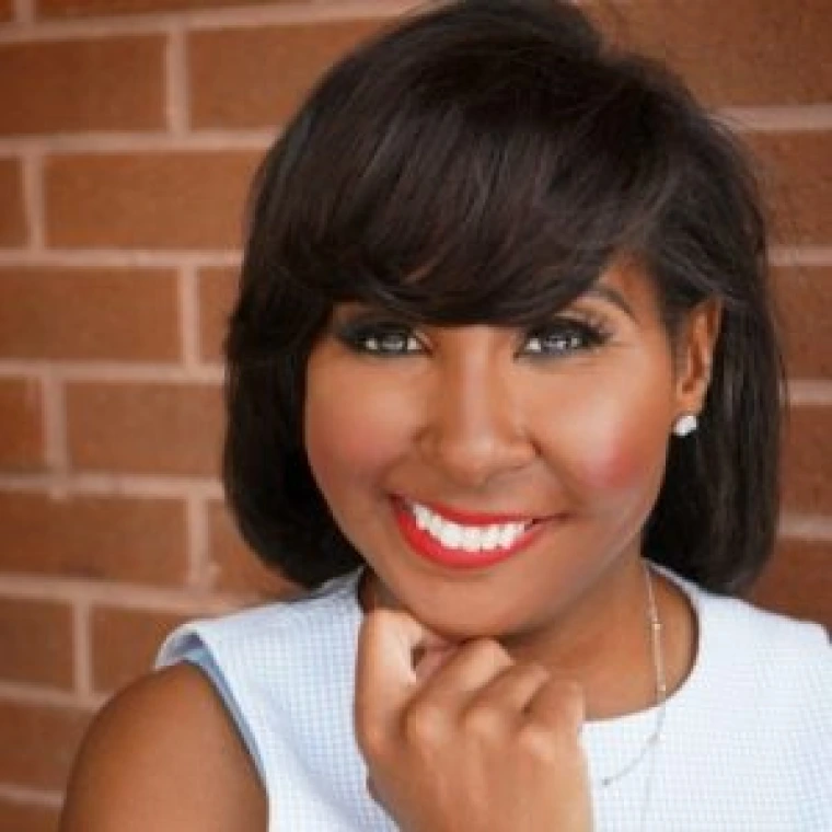 Black woman with medium-dark brown hair. She is wearing a sleeveless blue and white checkered top, with a silver necklace, wearing red lipstick smiling.
