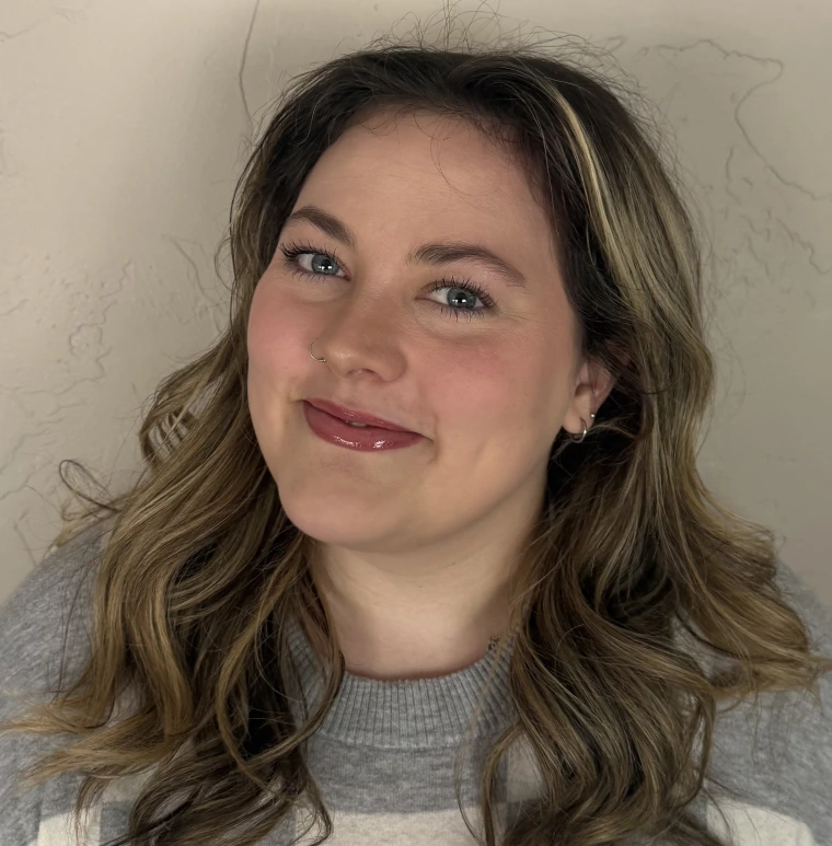 A woman against a white wall smiling. She has brown hair with blonde highlights and light pink lip-gloss on she also has blue eyes. She is wearing a white and grey sweater. 