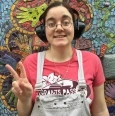 Photo of Abby, a woman with pulled-back brown hair, pale skin, wearing glasses, black headphones, a pink shirt, and a white apron. She is smiling and throwing a peace sign.