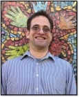 Photo of Bradley, a man with dark brown hair pale skin, and chin stubble. He is wearing glasses and a blue and white striped button down shirt in front of a mosaic mural of a flower.