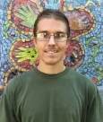 Image of Cody, a man with pulled back brown hair, tan skin, and glasses wearing a green T-shirt and smiling in front of a mosaic mural.