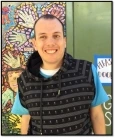 Image of Jack, a man with short brown hair waering a black sweater vest over a blue T-shirt, smiling in front of a mosaic mural.
