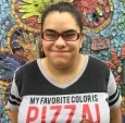 Image of Jennessa, a woman with brown hair in a ponytail, red-framed glasses, an a black and white shirt that says "Pizza" on it, smiling in front of a mosaic mural.