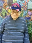 Image of Joe, an elderly man with glasses and wearing a blue and red University of Arizona cap and a black and white wtriped shirt, smiling in front of a mosaic mural.