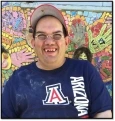 Image of Joey, a man with brown hair, wearing a red baseball cap and a navy blue U of A shirt, smiling in front of a mosaic mural.