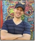 Image of Jon, a man wearing a black baseball cap and wearing a navy and black striped shirt, smiling in front of a mosaic mural.