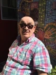 Image of Marco, a bald man wearing sunglasses and a pink and blue plaid shirt, smiling in front of a mosaic mural.