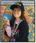 Image of Monica, a woman with long brown hair wearing a black hat and a black long-sleeved shirt, holding a blue ball with a face and a mustache on it, smiling in front of a mosaic mural.