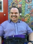 Image of Paul, a man with short brown hair and a mustache, wearing a blue button-down collared shirt, smiling in front of a mosaic mural.