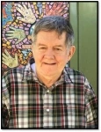 Image of Ricky, a man with short gray hair and a black, white, and red flannel shirt, smiling in front of a mosaic mural.