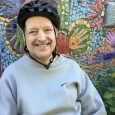 Image of Ron, an elderly man wearing a black bike helment and a grey sweatshirt, smiling in front of a mosaic mural.