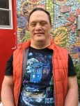 Image of Scott, a man with short brown hair wearing a black T-shirt and  red puffer vest, smiling in front of a mosaic mural.