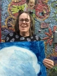 Image of Tina, a woman with shoulder-length brown hair and glasses, wearing a black and white shirt and holding up a blue and white painting while smiling and standing in front of a mosaic mural.