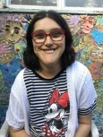 Image of Jen, a woman with short dark brown hair and square, red-framed glasses, wearing a black and white striped shirt with an image of Minnie Mouse on it, smiling in front of a mosaic mural.