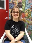 Image of Vicky, a girl with short brown hair and wispy bangs, wearing glasses and a black shirt with white stars, smiling in front of a mosaic mural.