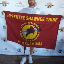 Native man with a prosthetic leg holding a red Absentee Shawnee Tribe flag