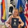 Reagan, a white woman in a pink suit and who sits in a power wheelchair, holds up a folder that says MDA Advocacy on the cover. Reagan sits in front of several inclusive flags, alongside the state of Wisconsin flag and the American flag. Reagan smiles at the camera.