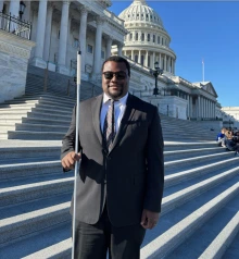 Photo of Jordan Moon, a black man wearing sunglasses and a suit and tie. He is holding a cane and standing in front of the U.S. Capitol Building.